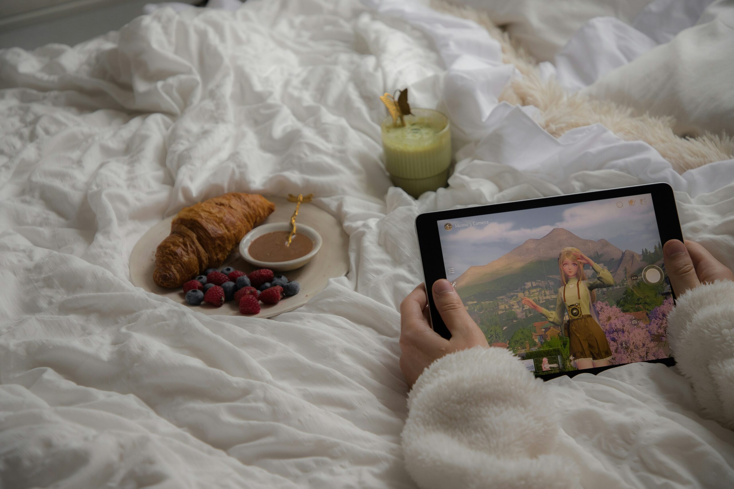 Hands holding a tablet playing 'Infinity Nikki', with a relaxing breakfast in bed situation featuring a matcha latte & croissant in the background.