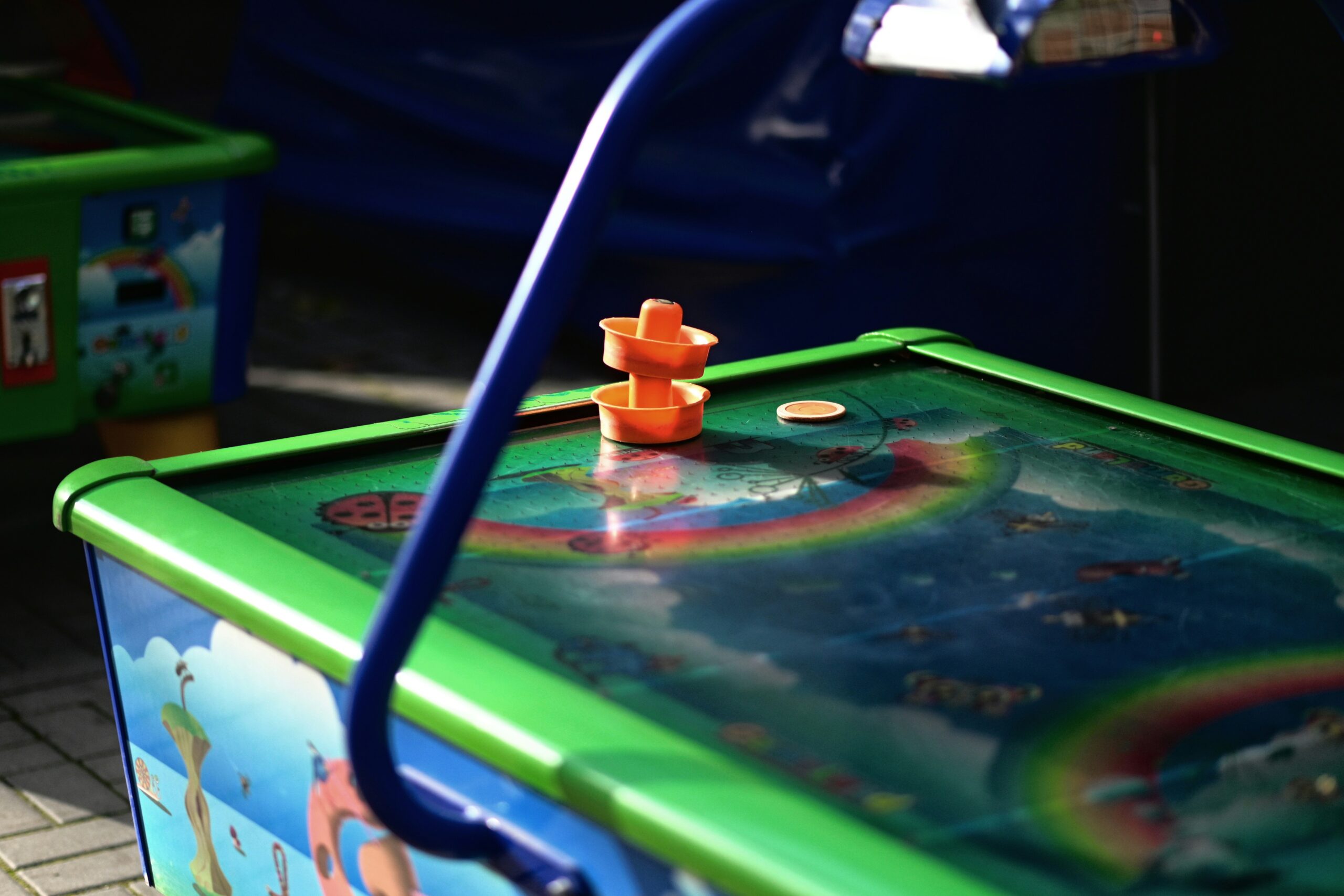 This close-up shot focuses on a small, vibrant air hockey table, likely designed for children. The table's surface features a colorful, cartoon-like underwater scene with fish and a rainbow. An orange paddle and a small, white puck are resting on the playing surface. The table's frame is a bright green, contrasting with the blue legs and other elements visible in the background.